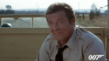 Movie gif. Roger Moore as James Bond in Octopussy. He's riding in the back of a truck and he gives an unabashed smirk and shrug of his shoulders as he rides away. 