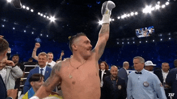Video gif. Boxer Oleksander Usyk raises his arms victoriously, bouncing up and down with a proud expression as fans in the background watch in awe and do the same. 