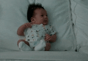 Video gif. A tiny baby sits propped up on a couch, then sneezes and topples over.