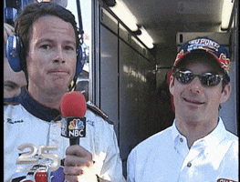 Sports gif. Jeff Gordon stands next to a man with a microphone up his face. We look at Jeff who smiles and crosses his fingers. 