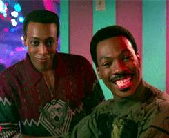 Movie gif. Eddie Murphy as Randy and Arsenio Hall as Semmi in Coming to America. They're at a club and they look at someone eagerly, with wide grins filling their faces as they lean in to hear better.