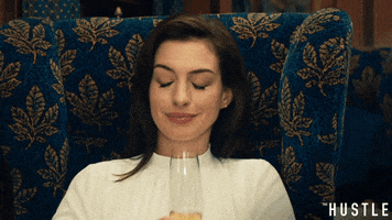 Movie gif. Anne Hathaway as Josephine in The Hustle glances down while smiling and tipping her champagne glass forward.