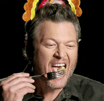 TV gif. In a scene from The Voice, we see a closeup of Blake Shelton wearing a Thanksgiving turkey decoration on his head. He has a complicated expression, not quite thrilled but not unhappy, as he eats a bit of food from a fork.