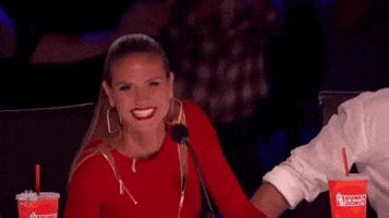 TV gif. Heidi Klum on America's Got Talent judges' panel, smiling and waving with both hands at us, as Mel B creeps in from the side with an expression of shock on her face.