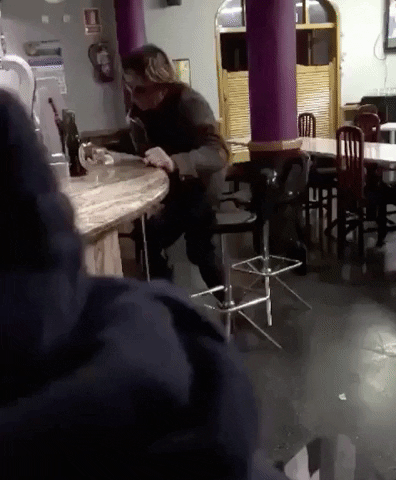 Video gif. A drunk man attempts to sit on a barstool but stumbles, falling head-first onto the floor.