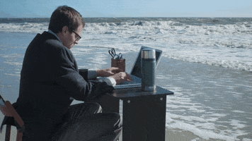 Video gif. A man has his office set up at the beach and he's sitting in a chair on the shoreline, feet touching the waves, while frantically typing on his laptop.