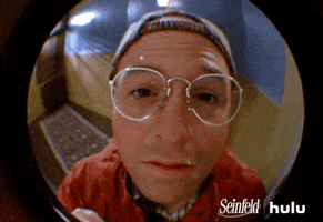 Seinfeld gif. Jason Alexander as George peers into Jerry Seinfeld's peephole from outside of his apartment. He waves hello and offers a weak smile as he waits to be let in. 