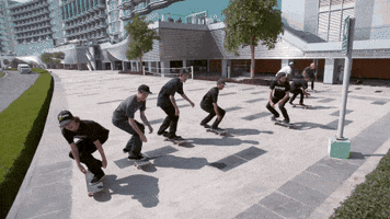 Sports gif. Eight skateboarders jump over a large hedge and land on the other side in unison. The apex of their jump is in slow motion, then resuming normal speed as they land and zoom away.