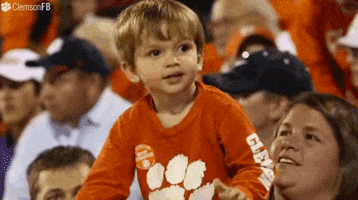 Sports gif. Toddler in the crowd at a Clemson Tigers game is standing up and clapping.