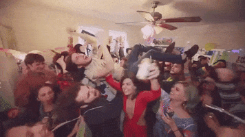 Video gif. Rowdy house party with people drinking, screaming, and holding up an ecstatically happy young man as he crowd surfs on his back. 