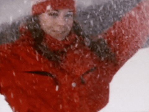 Music video gif. Mariah Carey in the All I want For Christmas is You Music Video is out in the snow with a red winter coat and hat on. She looks at us, smiling, and tossing snow in the air. 