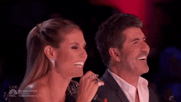 Reality TV gif. Heidi Klum and Simon Cowell on America's Got Talent sit at the judge's table laughing, Heidi throwing her head back and opening her mouth wide in laughter. Simon is laughing too, but his face is less expressive...wonder why?