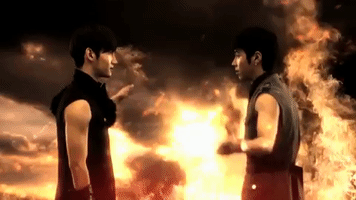 Celebrity gif. Standing in front of an epic, explosive flaming background, Yunho and Changmin from TVXQ high five each other.