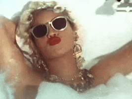 Celebrity gif. Beyonce sits in a bubbly bathtub with golden jewelry around the neck and blacked out golden sunglasses on. Her face. She kisses the air with her red lips. 