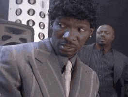 TV gif. Charlie Murphy on the Chappelle Show, dressed in a striped gray suit with a black curly-hair piece, looks down over a conference table skeptically, then cracks up emphatically and high-fives the other men in the room.