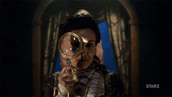 TV gif. Woman dressed in an extravagant lacy gown from the 1800s holds up a large gold magnifying glass and peers through it as she slowly turns from one side to the other.