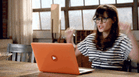 TV gif. Zooey Deschanel as Jessica Day on New Girl sits in front of her laptop at a table. She looks at the screen and raises her fists up in excitement.