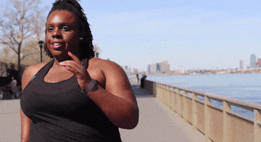 Video gif. Heavyset woman runs happily along a trail overlooking the water and a sunny cityscape.