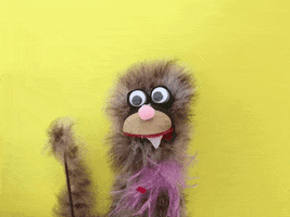 Video gif. A fuzzy puppet with different shades of brown hair, has large bobble eyes. One pointed tooth sticks out from under its tan snout. A stick holds up one of the puppets hands. It's long arm waves quickly. 