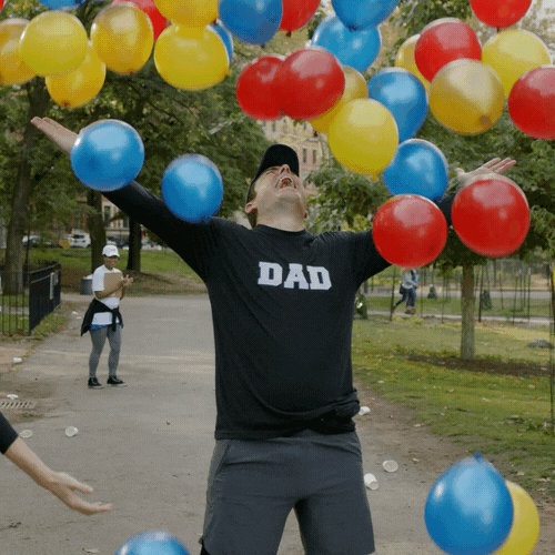 TV gif. Jason Jones as himself on The Detour is wearing a shirt that says, "Dad," is standing in the middle of a park with his arms outstretched. He leans back and has his face to the air and he yelps with joy as a bunch of balloons fall around him.