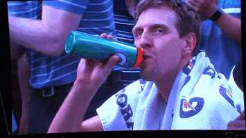 Sports gif. Dirk Nowitzki from the Dallas Mavericks is on the bench drinking water and he gets caught on the jumbotron. He raises a hand and waves at the crowd.