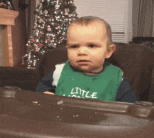 Video gif. Baby in a green bib sits in a high chair raising its eyebrows and smirking expressively as it holds up a pointed finger.
