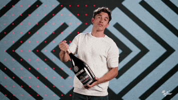 Sports gif. Marc Marquez, a motorcycle racer, opens a bottle of champagne and it sprays into the air with force. Some of the champagne lands on him. 