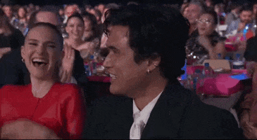Celebrity gif. Charles Melton slightly, shyly dips his head into his fingertips momentarily looking away from the awards show crowd before pulling his head up again with a big smile recovering from his moment of embarrassment. Natalie Portman sits next to him rocking back and forth with glee as she looks at Charles and claps her hands.