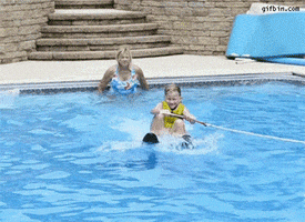 Video gif. Girl crouches on water skis as she's pulled slowly through a pool. She looks to the side as a dog off screen leaps onto her, plunging them both into the water.