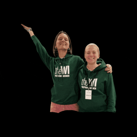 Photo gif. Two women wearing matching green "Women's Institute" hoodies smile and stand next to each other with their arms around each other's shoulders. One of the women has her arm raised in the air, looking proud, as the photo changes back and forth between mirrored images. 