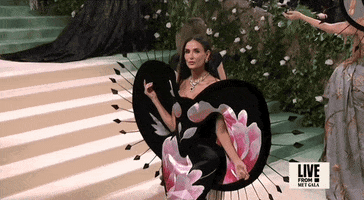 Met Gala 2024 gif. Demi Moore holds one hand delicately up to her black and floral Harris Reed velvet gown that features a double duchess silk panel adorned with massive pink peonies. Large black petals arc out from her neckline down to her hips in a semi-heart shaped pattern. The petals are edged with black thorns with arrowheads at the end.