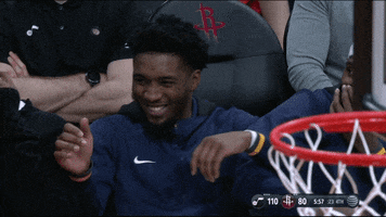 Sports gif. Donovan Mitchell from the Utah Jazz is cracking up on the bench with his teammates. He laughs so hard that he nearly falls out of his seat, and he lays against his teammates who are all laughing just as hard.