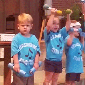 Video gif. Young children perform on a church stage holding barbell props except one kid, who stays still and stares at us, annoyed.