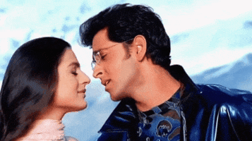 Celebrity gif. Hrithik Roshan slowly kisses each of a woman’s closed eyes as she leans in and smiles.