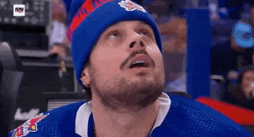 Sports gif. Auston Matthews from the Toronto Maple Leafs wears a beanie and sits on the sidelines of the NHL All-Star Game. He watches the screen above him before turning to the side, making a surprised expression towards his teammates.  
