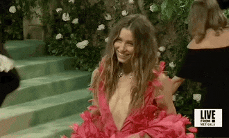 Met Gala 2024 gif. Closeup of the top of Jessica Biel's hot pink Tamara Ralph Couture gown with a plunging wide v-shaped neckline that falls to her mid-torso. Delicate hot pink petals flare out from the thin straps and throughout the bodice. Her hair is styled in a messy beach-style waves.