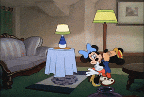 Cartoon gif. Mickey Mouse leans in to kiss Minnie as she pulls away shyly and a bunch of characters pop out from behind tables, lamps, and couches to surprise Mickey. Text, "Happy Birthday!'