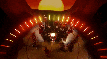 Oscars 2024 gif. Scott George and Osage singers perform 'Wahzhazhe (A Song For My People)' from Killers of the Flower Moon. We see the performance from overhead. An inner circle plays a glowing powow drum in unison while a group of singers in Indigenous wear dance around them and interact with each other. All of the performers are encircled by vertical lines of neon yellow and orange lights. 