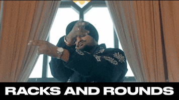 Celebrity gif. Indian rapper Sidhu Moose Wala, wearing a black turban, jacket, and gloves loads a magazine into a pistol while staring us down. Then he mimics recoil while firing a finger gun towards us. Text reads “Racks and Rounds,” occasionally glitching.