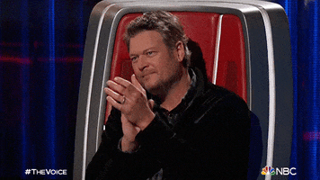 Reality TV gif. Blake Shelton sits as a judge on The Voice and applauds and smiles with a proud expression.