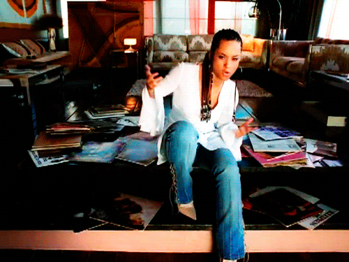 An animated gif of Alicia Keys sitting on a coffee table covered in books making a "what?" gesture