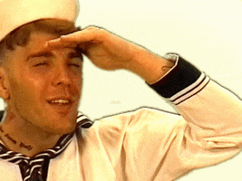 Video gif. A man wears a sailor outfit and hat. He cups his hand over his eyes like a visor and squints. 