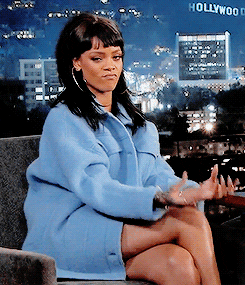 Celebrity gif. We see Rihanna in an armchair during an interview, wearing a light blue coat and large hoop earrings. Looking straight at us, she rubs her fingers and thumbs together as if to say "let's talk money."