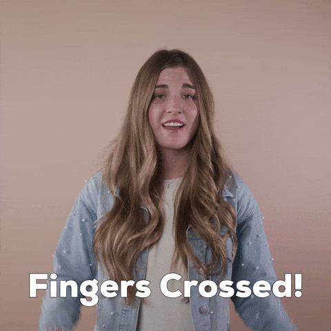 Reaction gif. A light-skinned woman with big eyes, shiny bronde hair, and cerebral palsy holds both hands up, fingers crossed, and with a nervous optimistic smile says, "Fingers crossed!"