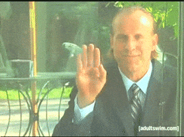 Video gif. Man with slicked back hair and dressed in a suit and tie waves mischievously through a window. A smirk plays upon his face as he stops waving, slowly spreads his fingers, and gives a thumbs up.