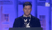 Video gif. Saturday Night Live's Colin Jost stands at a podium at the 2024 White House Correspondents' Dinner and takes a beat before acknowledging "Ok, I got one woo" while surveying the crowd.