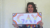 Video gif. A happy woman wearing a lei holds up a sign that reads, "GAY." She looks at the sign and does a fake expression of shock at what she's reading.
