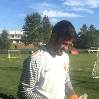 Sports gif. Alisson Becker of the AS Roma football team looks at us with a big smile and holds a yellow gloved thumbs up. He stands next to a practice field.