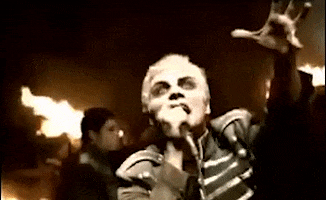 Music Video gif. Gerard Way in the My Chemical Romance Famous Last Words music video holds his hand out like he’s trying to reach towards something, and he angrily sings into the microphone in his hand. Behind him the dark landscape is on fire.