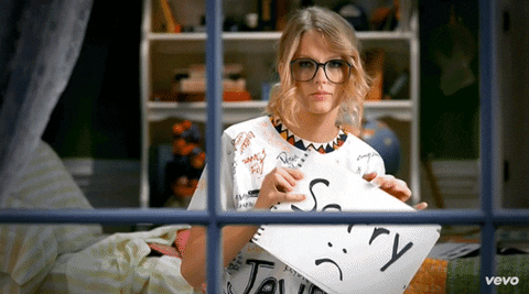 Music video gif. Taylor Swift in her video for You Belong with Me sits at a window facing us and holds up a note that says sorry as she gives a half frown.
